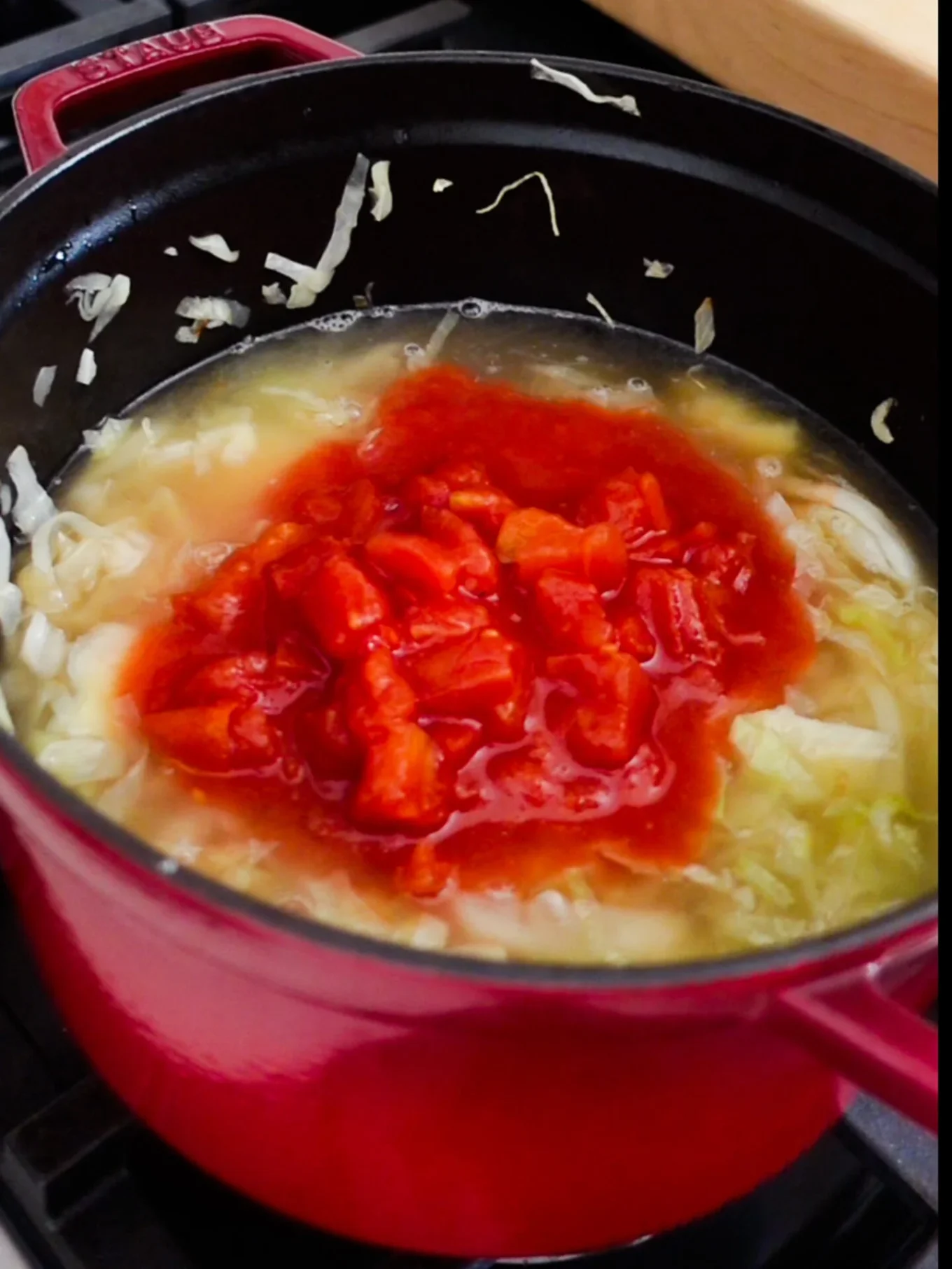 A red pot sits on a stovetop filled with chopped cabbage, broth, and a generous portion of diced tomatoes and tomato sauce being cooked together&mdash;steam rising as this Mexican Cabbage Soup, or Caldo de Repollo, simmers to perfection.