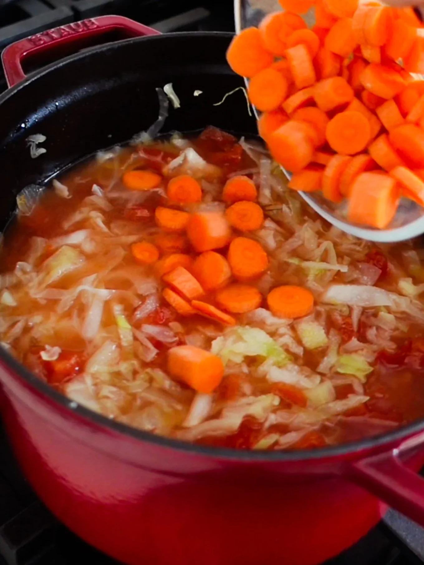 A close-up of a red pot on a stove shows Mexican Cabbage Soup simmering with chopped cabbage, tomatoes, and broth. Sliced carrots are being added, and the vibrant vegetables stand out in the steaming Caldo de Repollo.