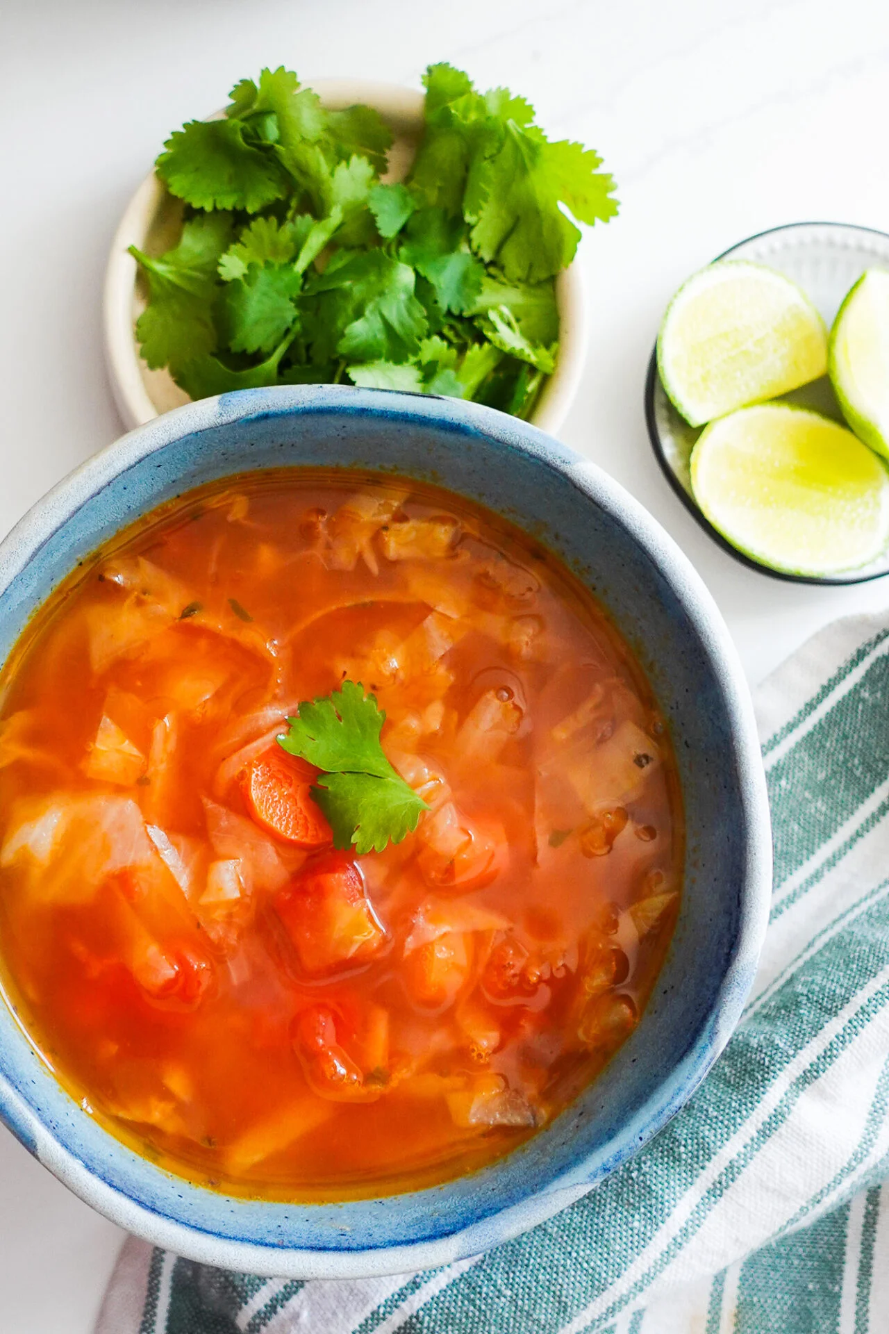 A bowl of Caldo de Repollo, or Mexican Cabbage Soup, with tomatoes and carrots sits on a white surface. Next to it are fresh cilantro, lime wedges, and a green striped cloth. The soup is garnished with a cilantro leaf.