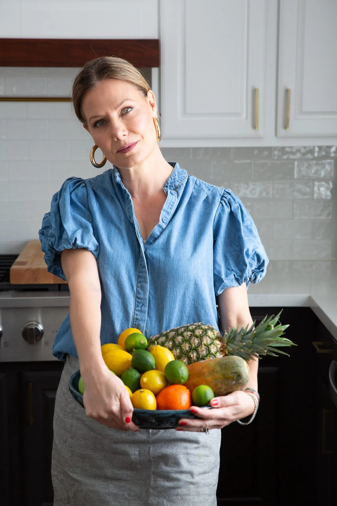 Alejandra Graf stands in a kitchen wearing a blue blouse and gray apron, holding a bowl of assorted fruits&mdash;lemons, limes, oranges, and pineapple. Her light hair is pulled back, with gold hoop earrings gleaming by the white cabinets and stove.