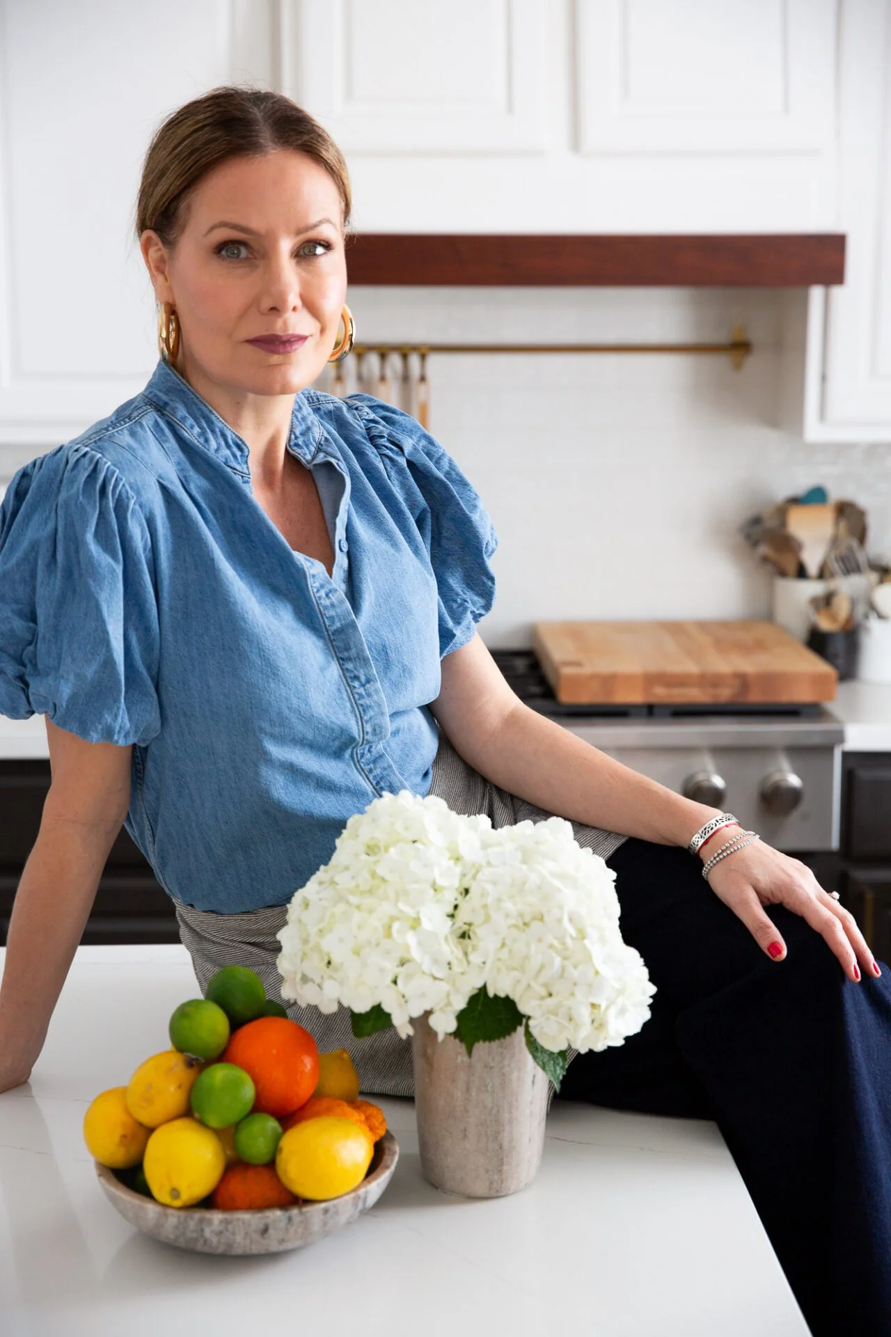 A woman in a blue puff-sleeve shirt sits on a white kitchen counter. In front of her are a silver vase with white hydrangeas and a bowl of lemons, limes, and oranges. The bright, modern kitchen has a wooden cutting board and utensils in the background.
