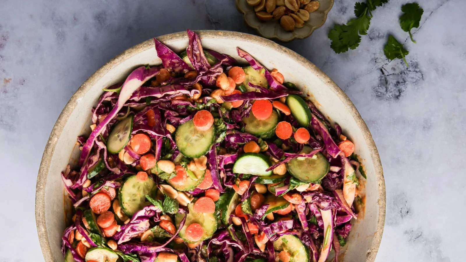 A bowl filled with colorful salad featuring sliced cucumbers, shredded purple cabbage, carrot rounds, chickpeas, and fresh herbs&mdash;perfect for effortless recipes and ideal as a vibrant addition to your next cookout. Peanuts and cilantro sit beside on a light, textured surface.