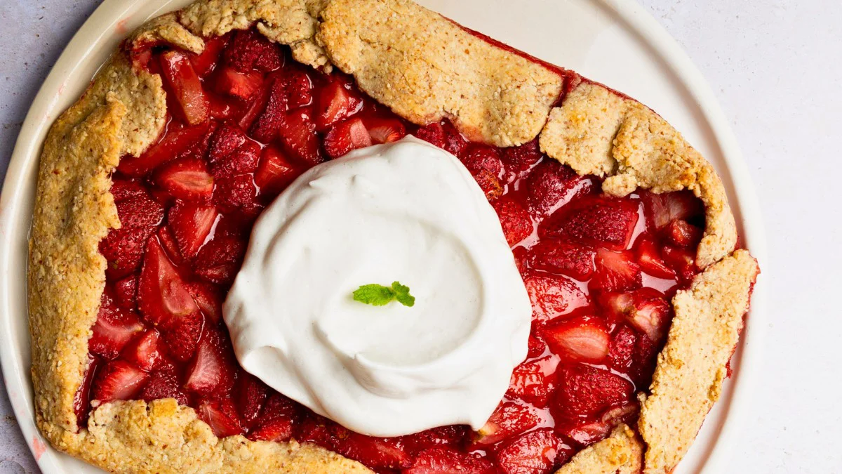 A rustic strawberry galette with a golden, flaky crust, filled with juicy strawberries&mdash;a delightful way to use summer produce. A large dollop of whipped cream topped with a small mint leaf sits in the center. The dessert is on a round, white plate.